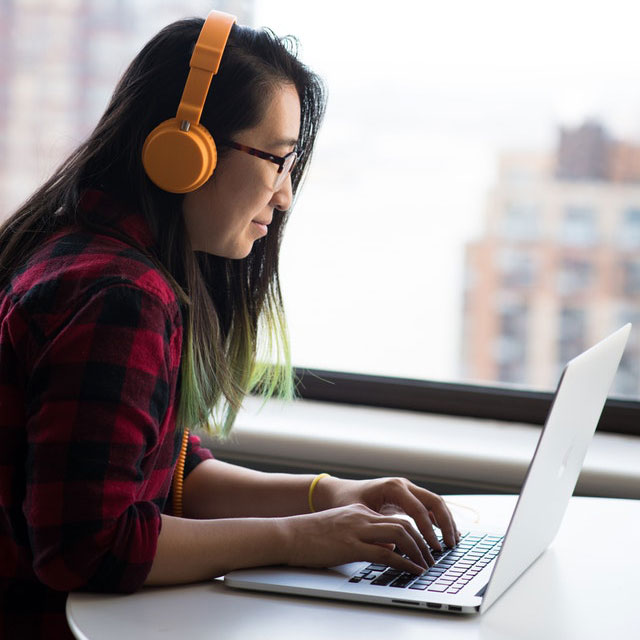 person with long dark hair wearing head phones looking at a laptop