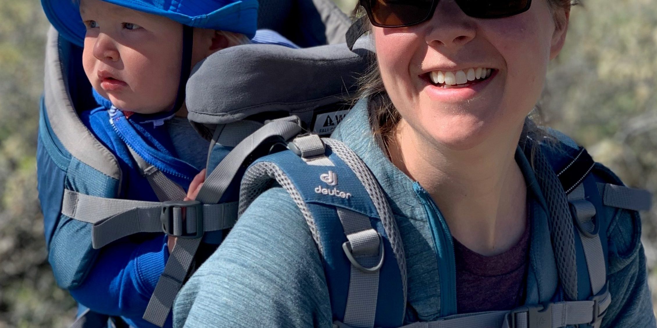 woman wearing yellow hat with baby in carrier on her back, baby is wearing blue hat