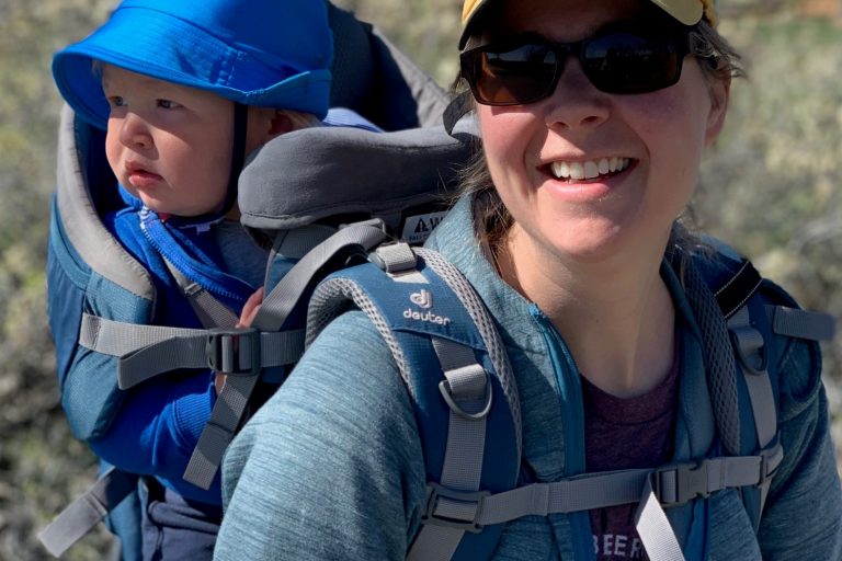 woman wearing yellow hat with baby in carrier on her back, baby is wearing blue hat