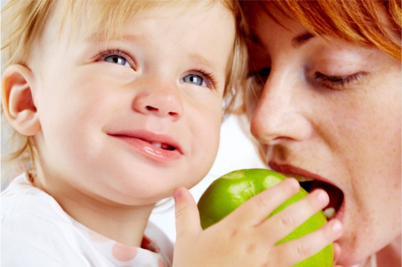 toddler with blonde hair holding a green apple older person leaning in to bite apple